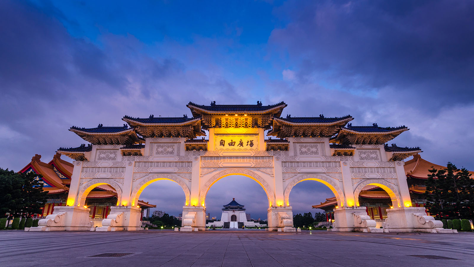 The front arch of Freedom Square is illuminated as night falls. In the distance Chiang Kai-shek Memorial Hall can be seen.