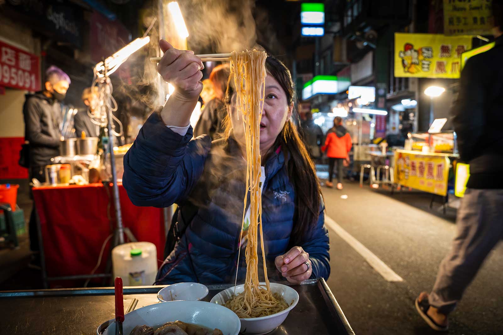 A bowl of steaming noodles in a night market.