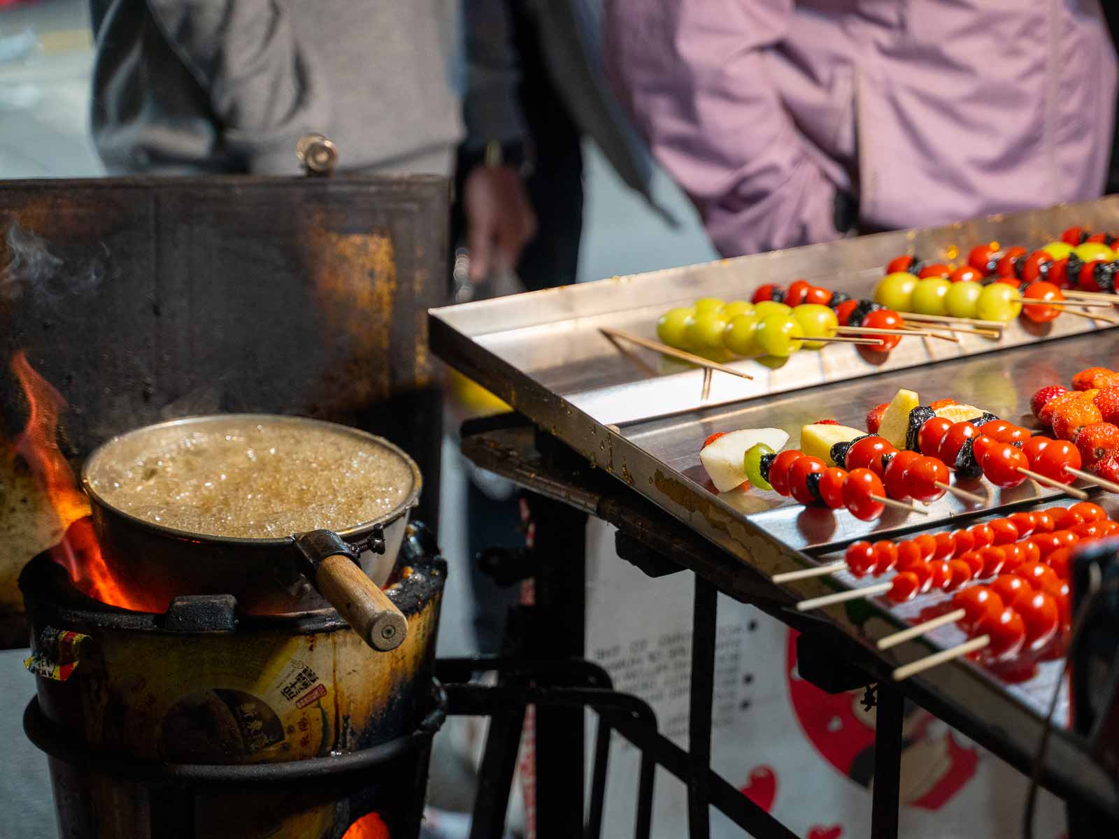 A boiling bowl of sugar-syrup sits next to fruit skewers.