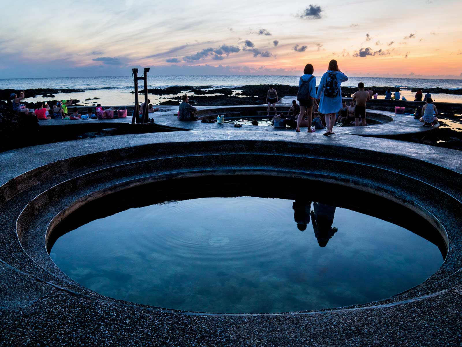 Travelers sit on the coast watching the sun, a circular hot spring pool appears in the foreground.