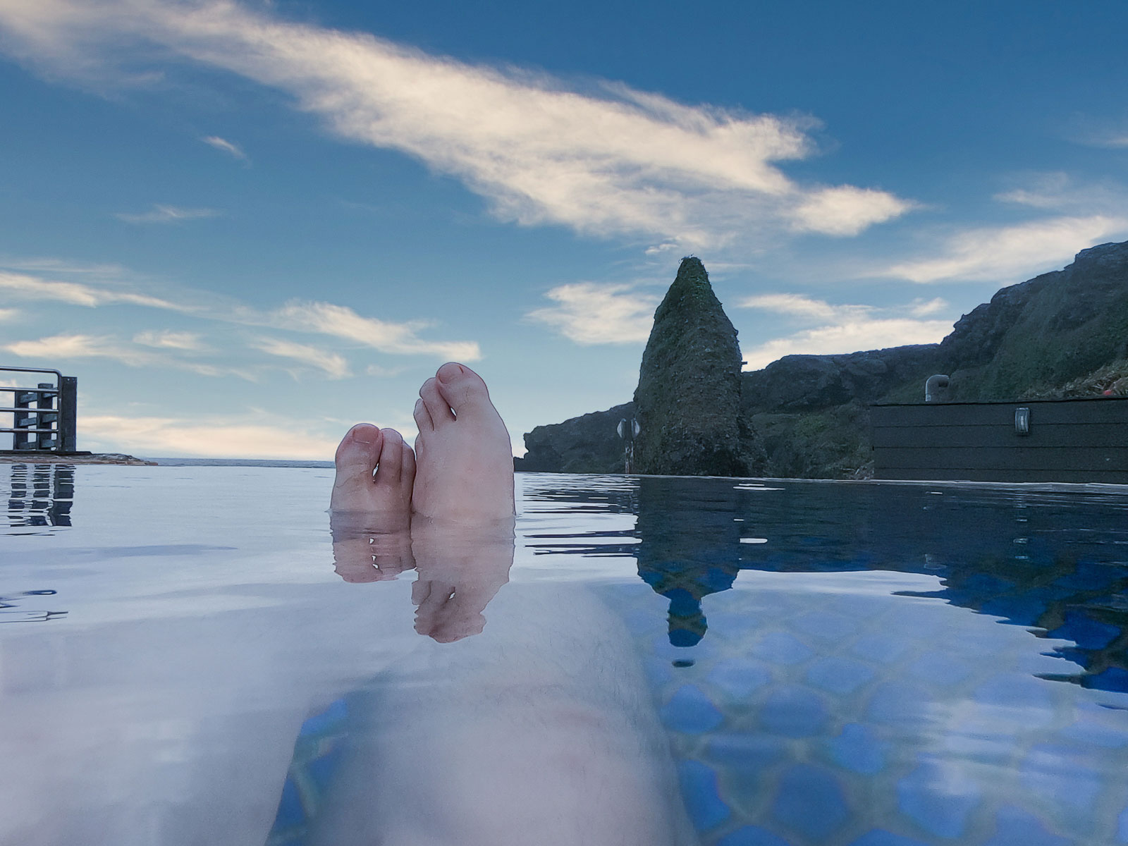 A first-person view from within the hot spring, a bather's feet emerge from the water and Green Island's coastal mountains can be seen in the background.