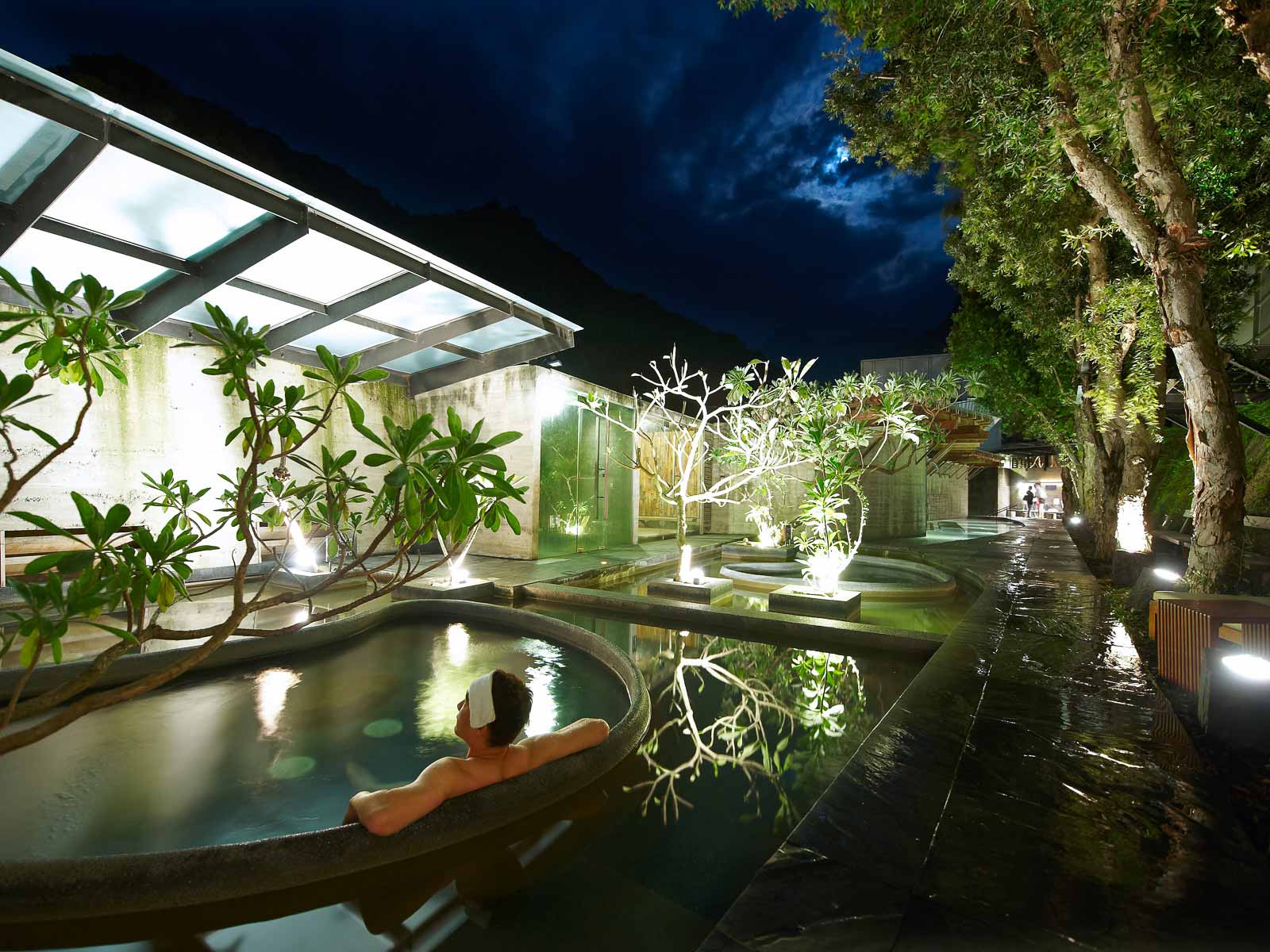 A bather soaks at night in a pool surrounded by sub-tropical plants.