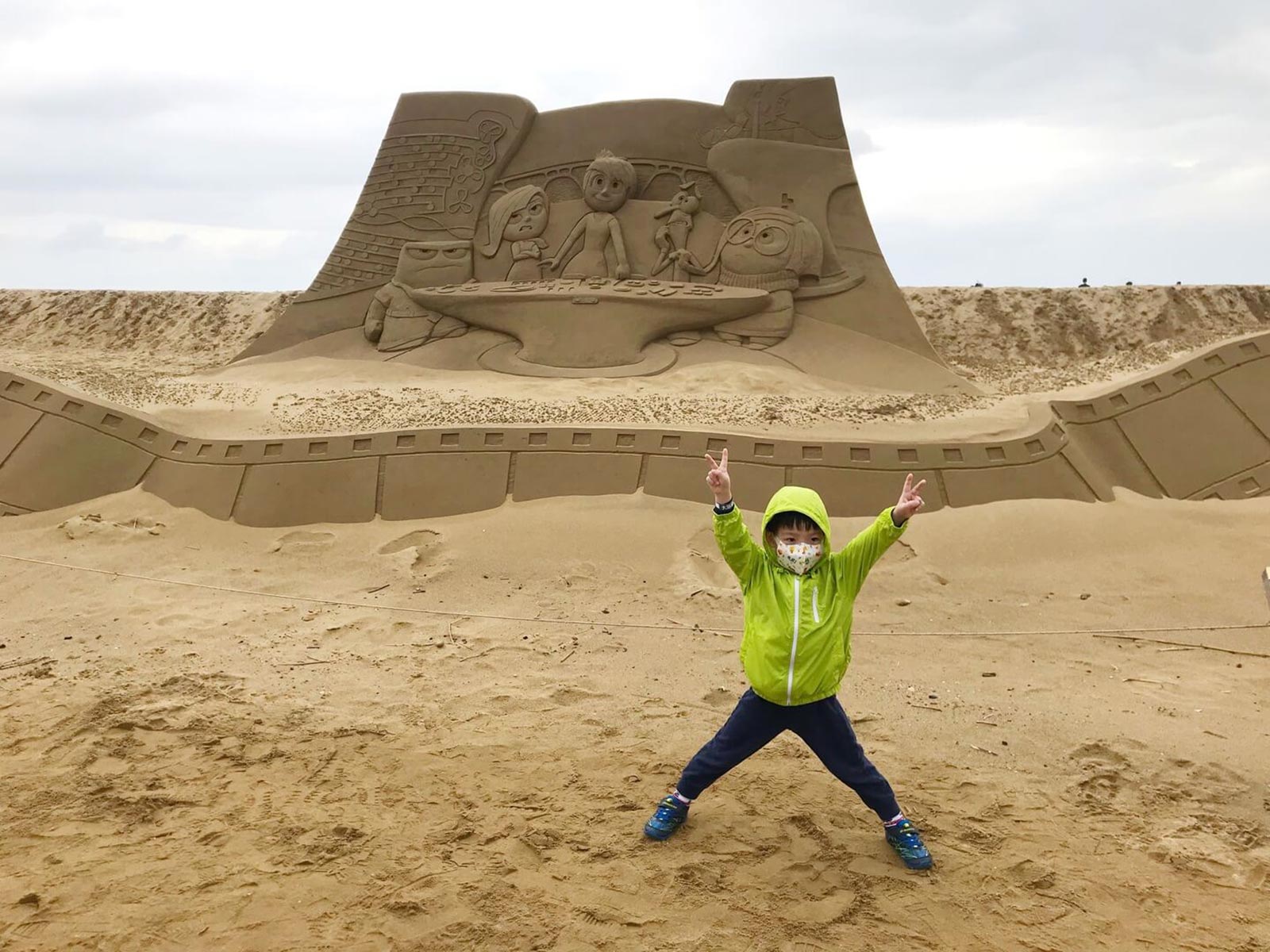 A youngster poses in front of a tall sand sculpture about the size of a car.