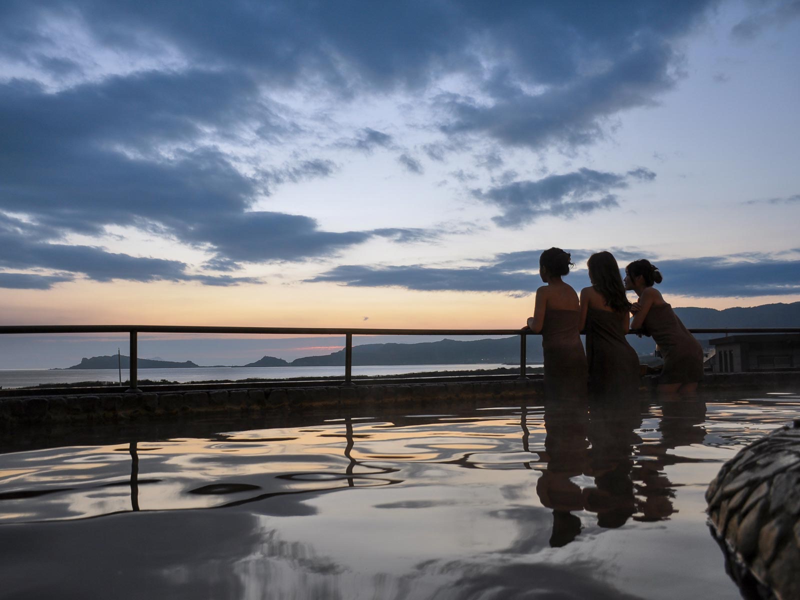 Bathers enjoy an outdoor hot spring while gazing at the ocean during twilight.
