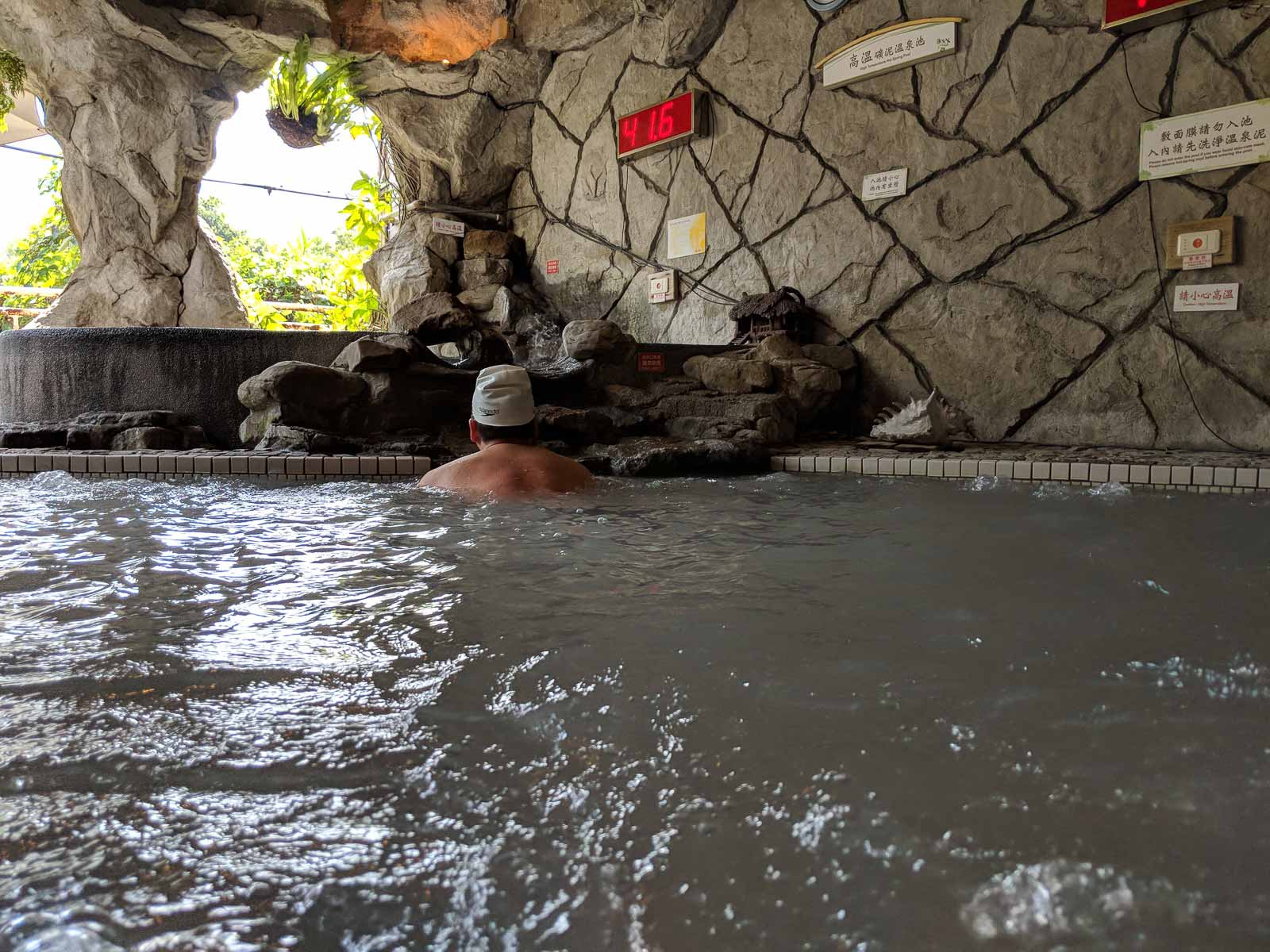 A hot spring bather soaking in muddy water, a thermometer reads 41.6 degrees Celsius.