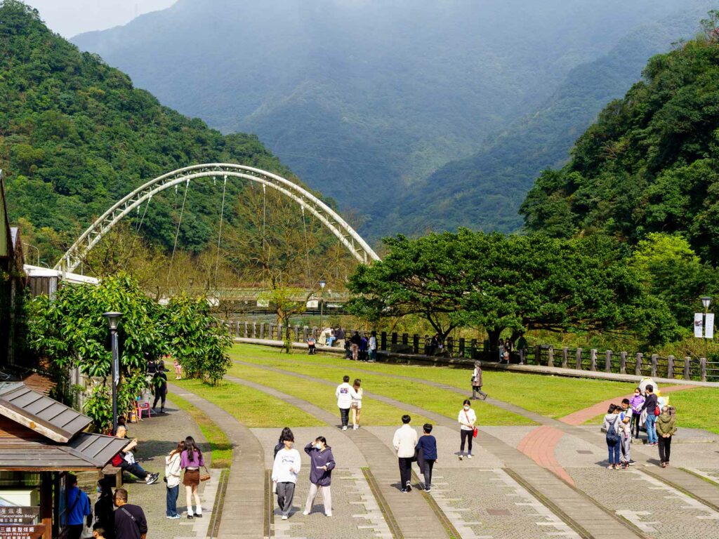 Jungle covered mountains rise behind a suspension bridge; tourists walk in a riverside park in the foreground.