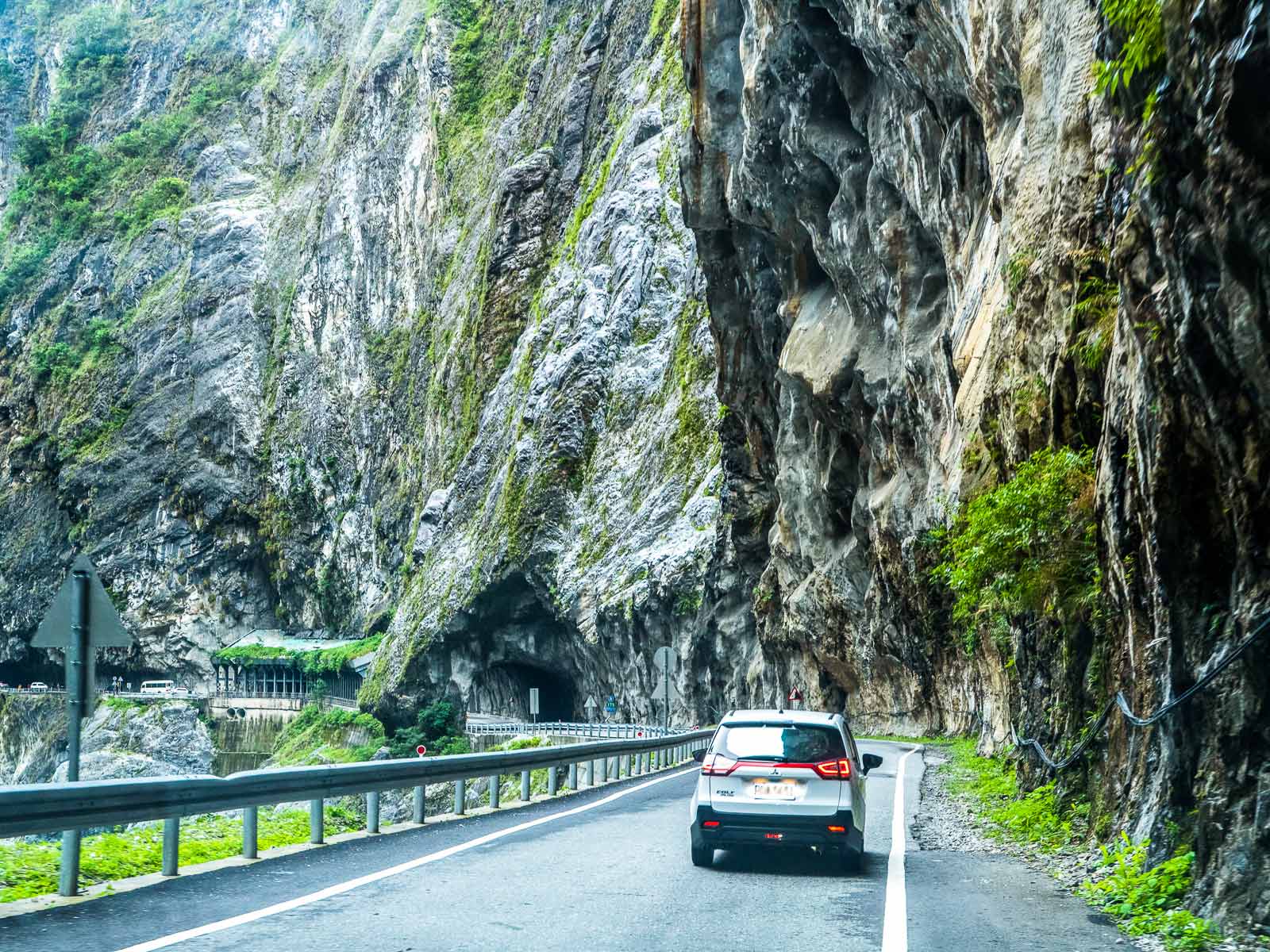 A car is seen driving underneath sheer cliffs with tunnels in the distance.