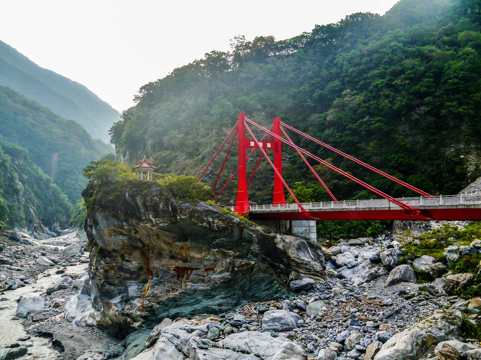 The colorful red Cimu Bridge crosses over a tributary of the Taroko River; a small shrine can be seen on a rocky outcrop.