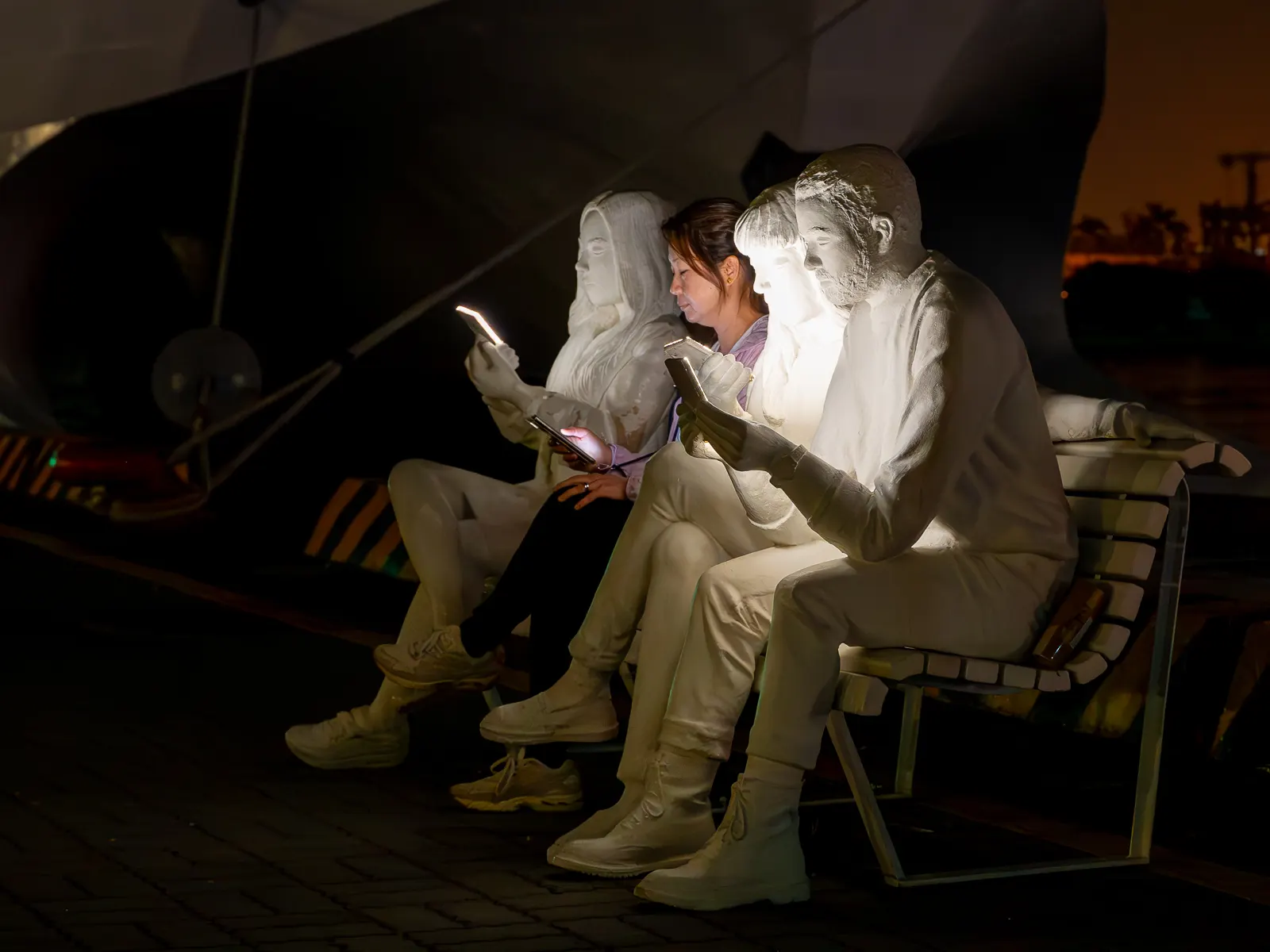 At night, a sculpture composed of statues sitting on a bench holding cell phones is illuminated by glowing screens.