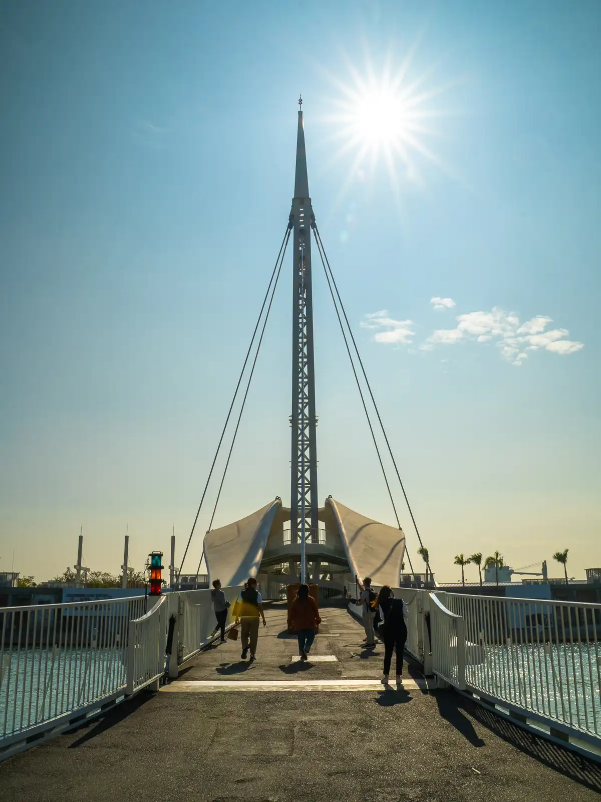 The sun shines overhead as tourists cross the bridge, which features tall railings on either side for protection.