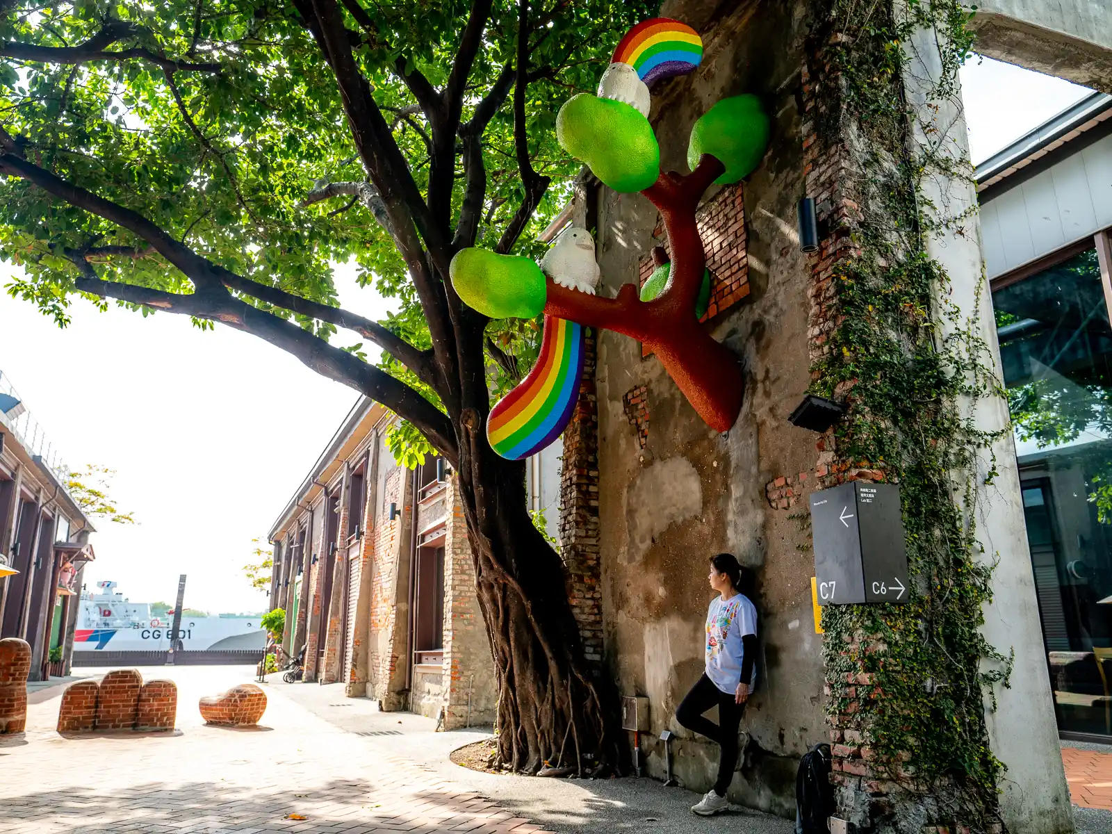 A sculpture of a tree with rainbows attached to it appears to grow right out of an old warehouse wall.