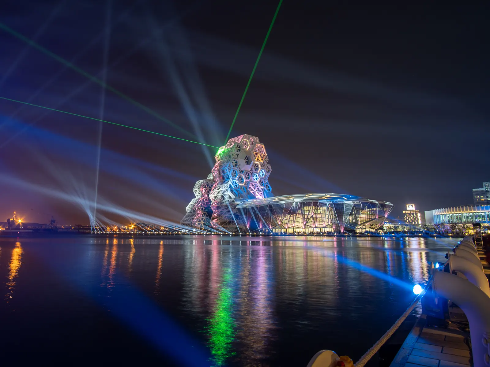 A wide shot of the Kaohsiung Music Center at night during a laser light show.