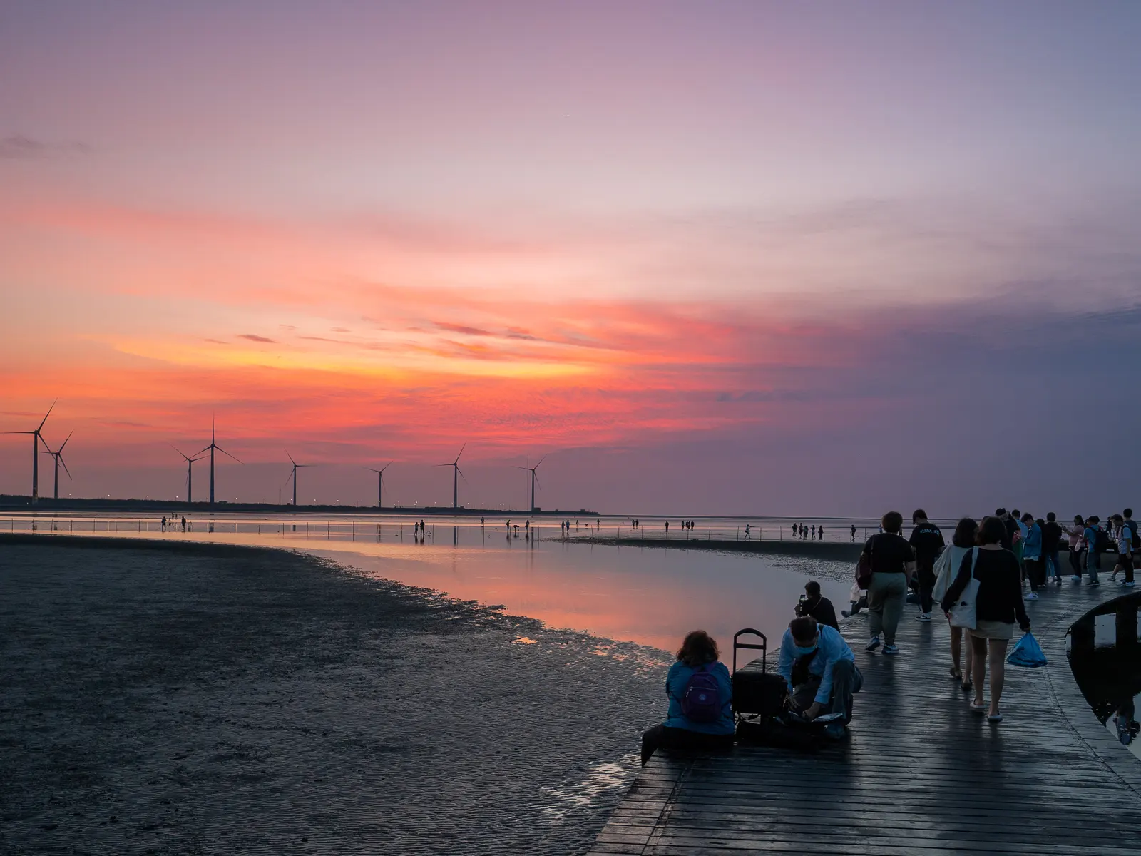 A group of tourists on a boardwalk watches the afterglow of the sunset.