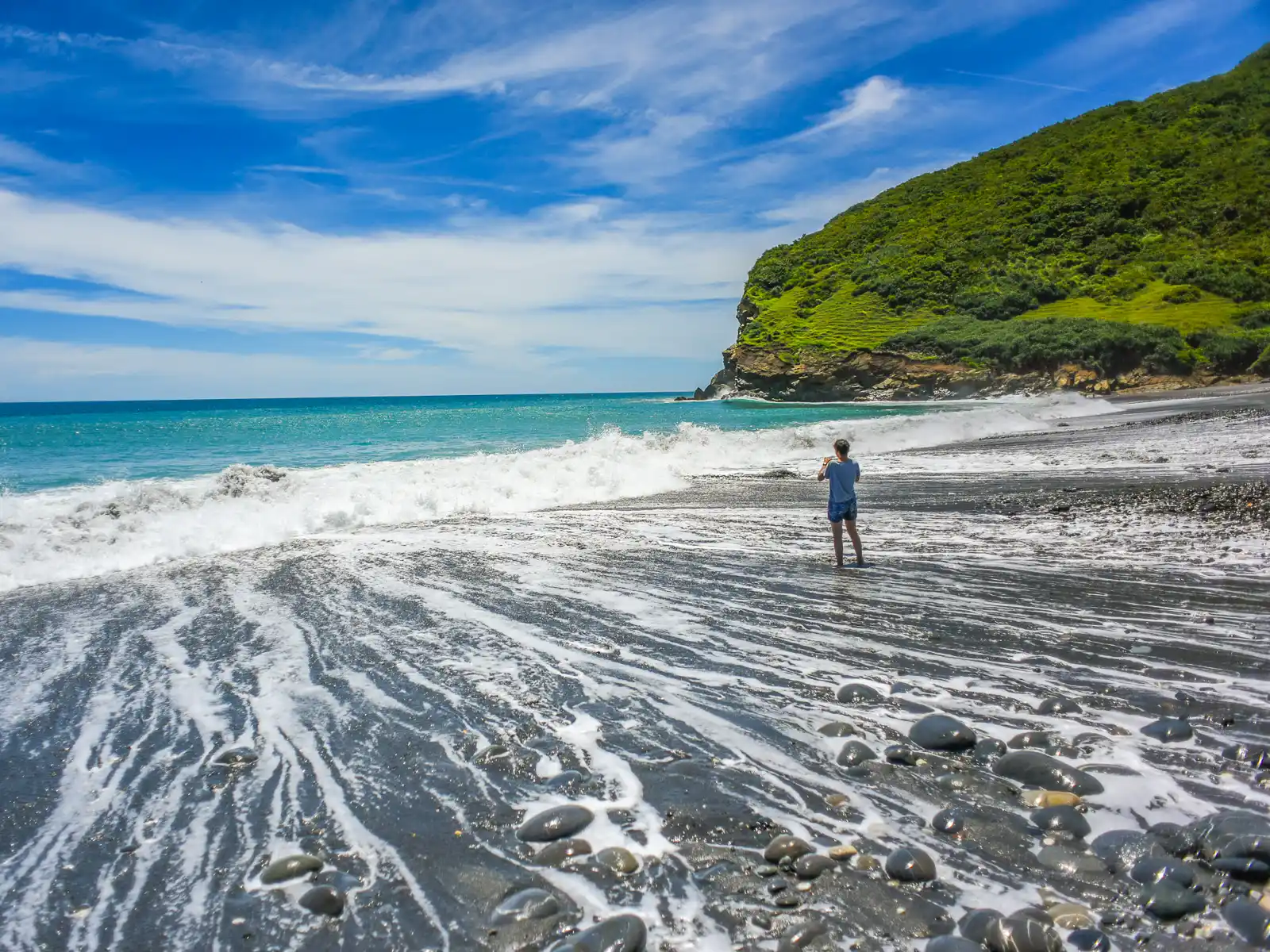 A tourist takes a photo as waves crash along a gently sloping black sand beach.