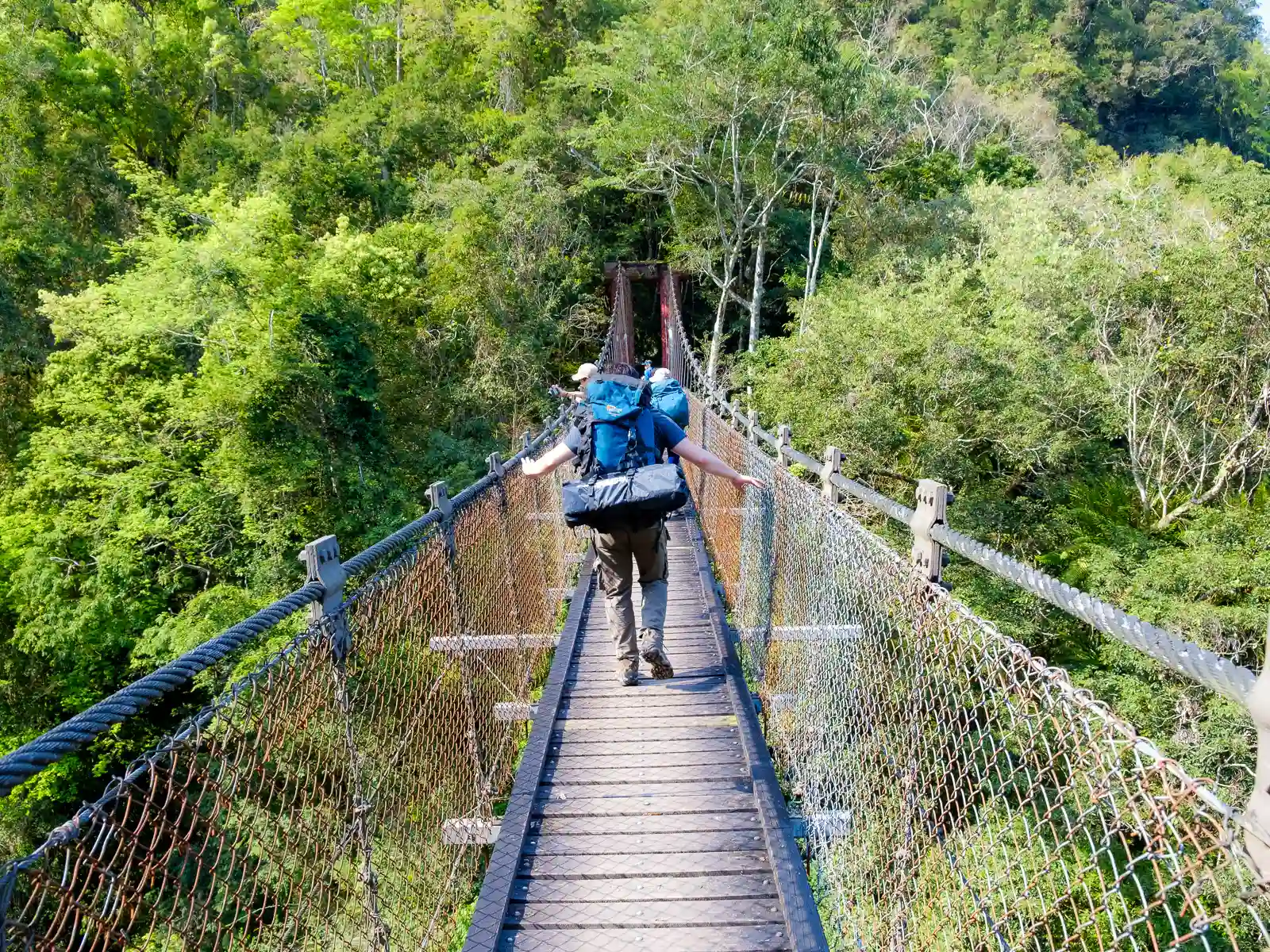 A hiker carrying a mountaineering pack crosses a suspension bridge.