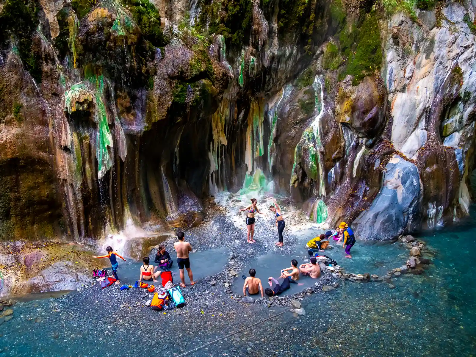 A group of tourists baths in hot spring pools below a colorful canyon wall.