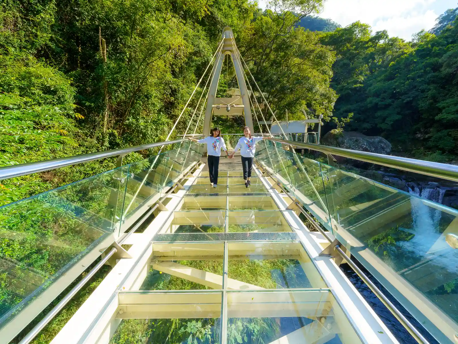 Two tourists hold hands as they walk down a glass-bottomed skywalk.