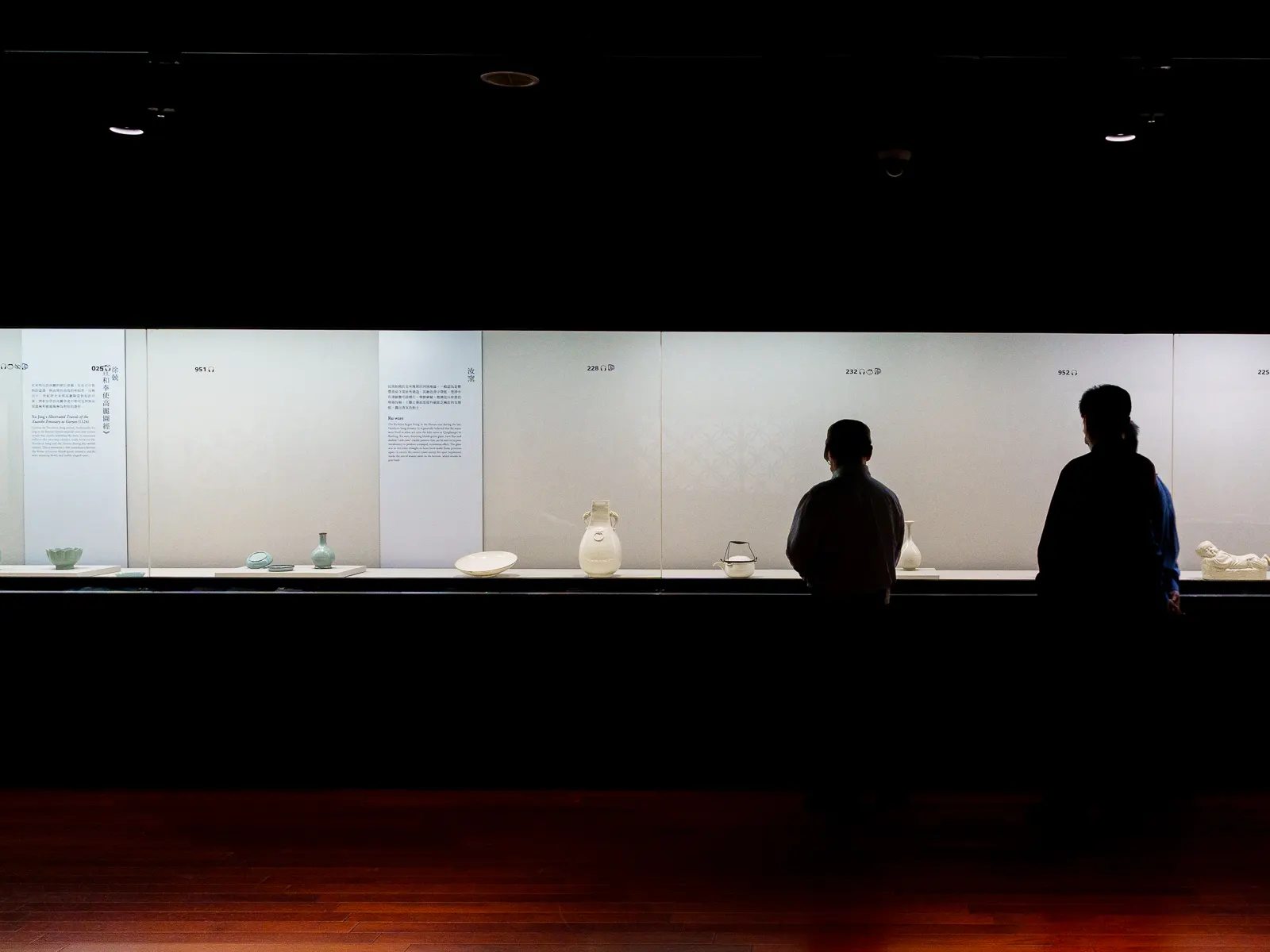 Two individuals observe white ceramic exhibits in a dimly lit museum gallery.
