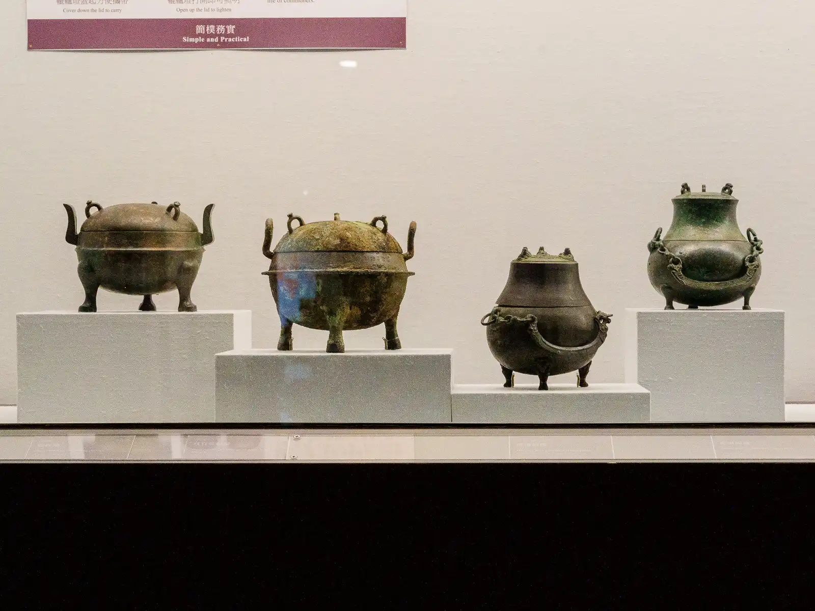 Four ancient metal pots are displayed on white pedestals against a white wall.