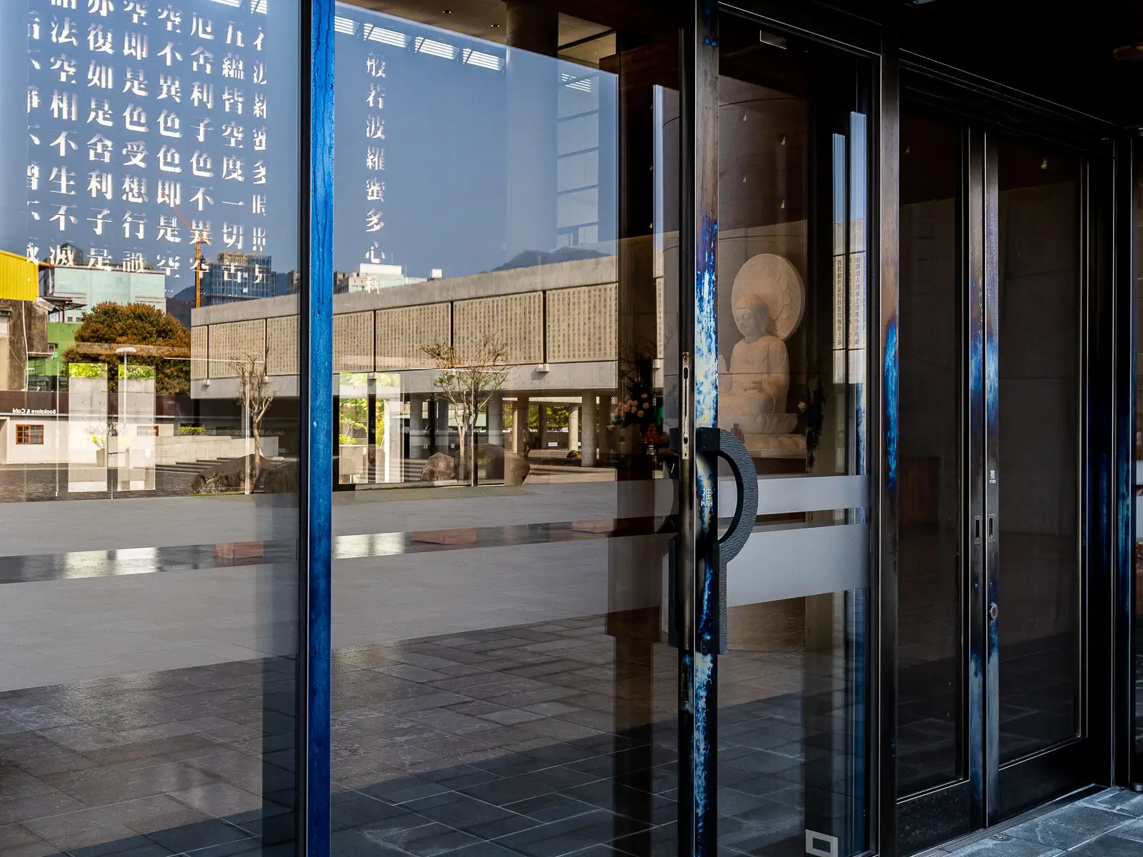 A Buddha statue is seen through glass doors, which also reflect architectural details of the temple's buildings.