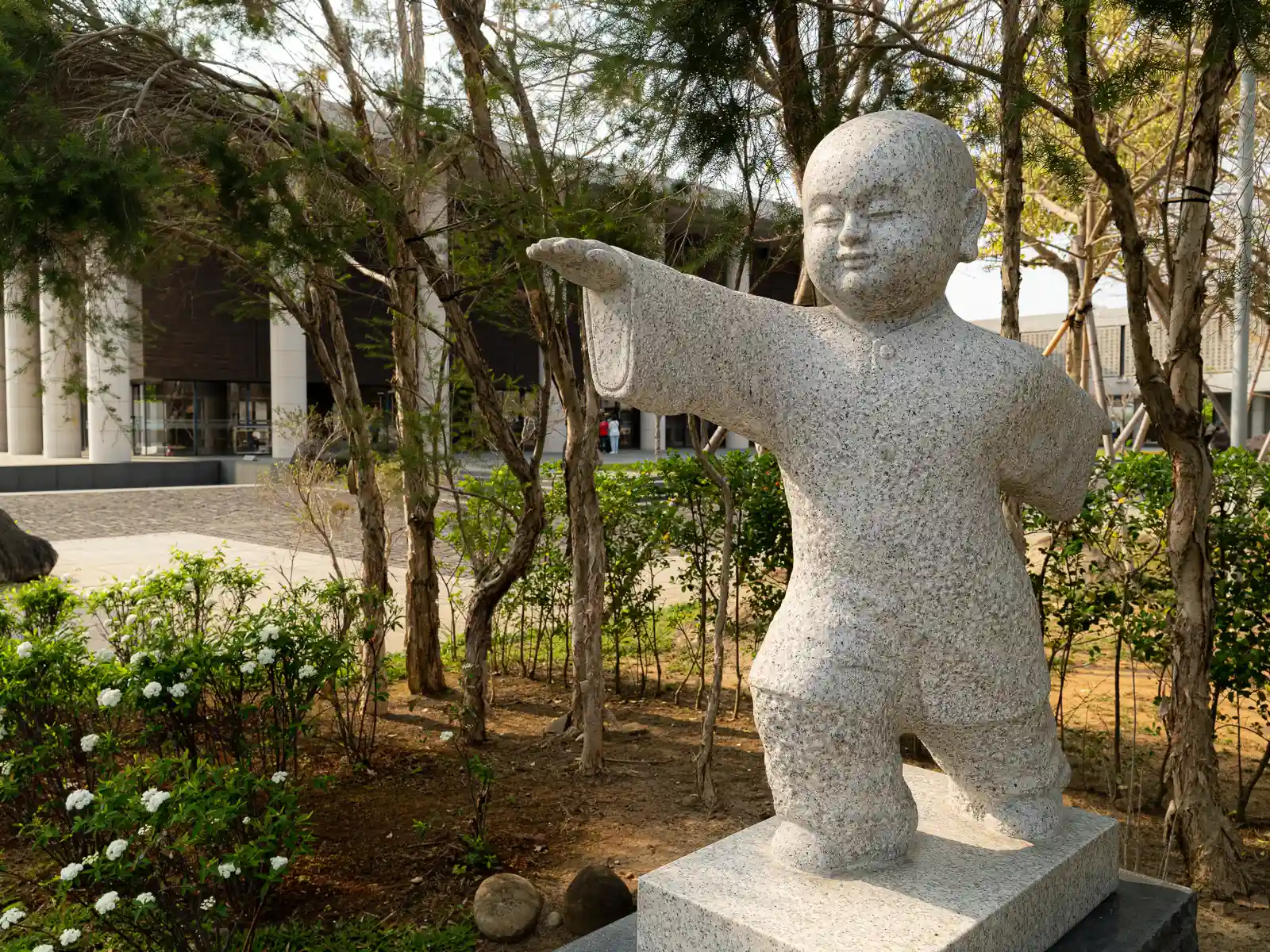 Granite statue of a child with an outstretched arm among trees and bushes.