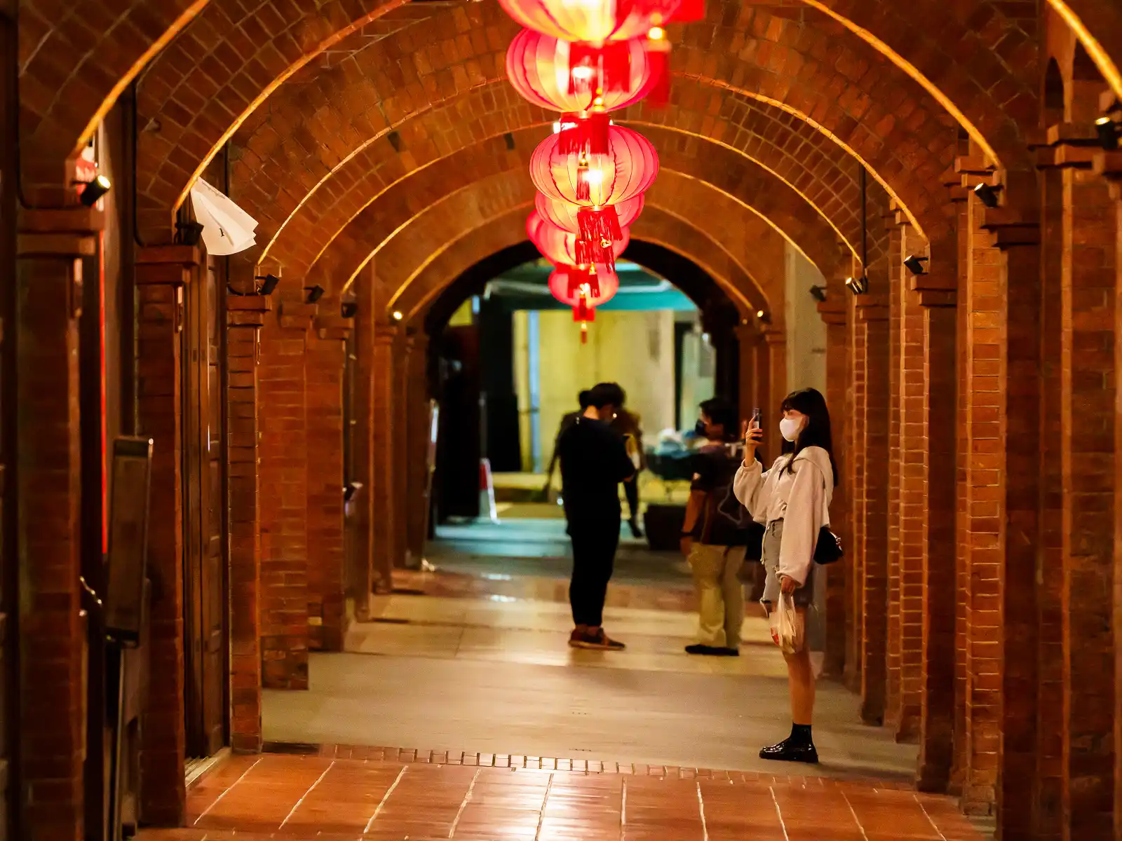 A red brick arcade decorated with red lanterns and a person taking a photo.