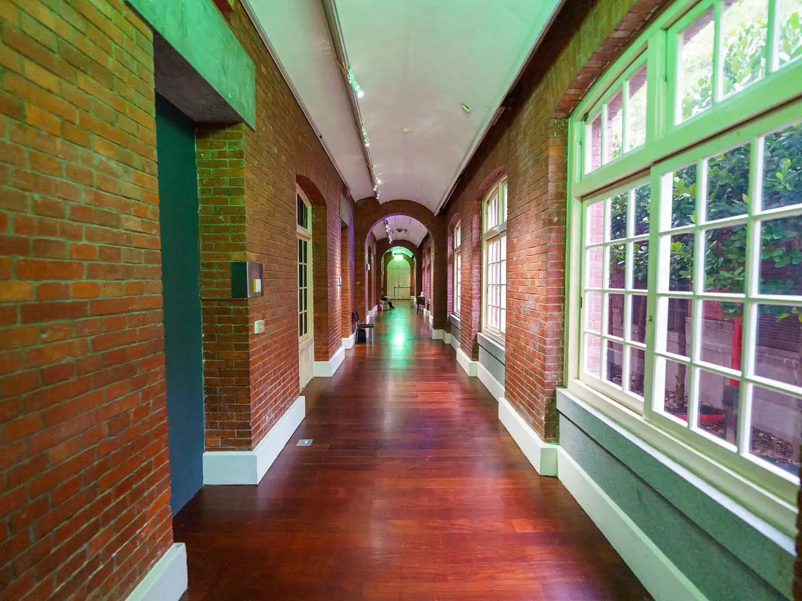 A hallway with wooden floors, red brick walls, and large windows on one side.