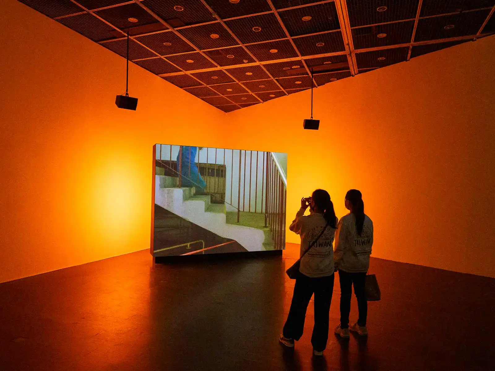 Two visitors watching a work of video art on a large screen set up in the middle of a orange room.