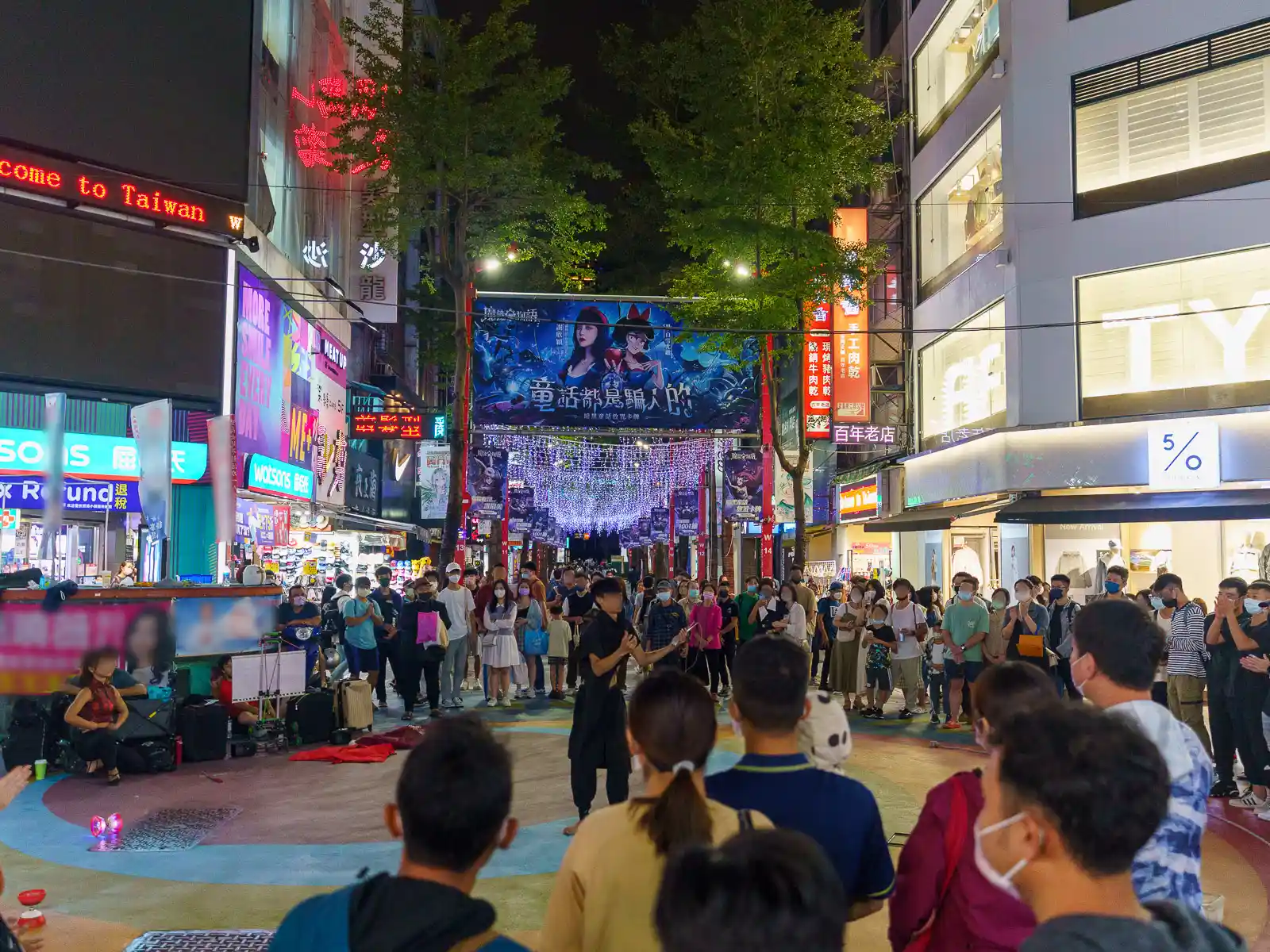 Night scene of a bustling street in Taiwan with pedestrians watching a street performer.