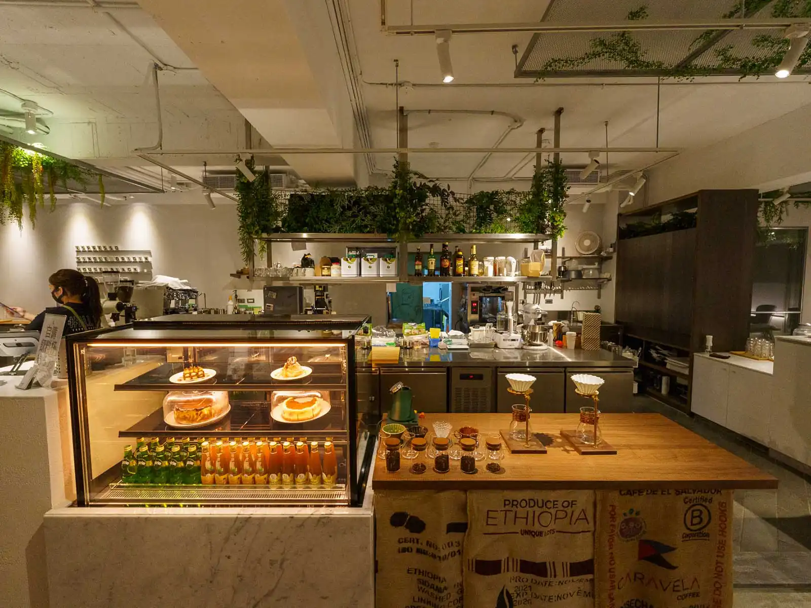 Modern cafe interior with desserts on display, hanging plants, and a well-equipped bar.