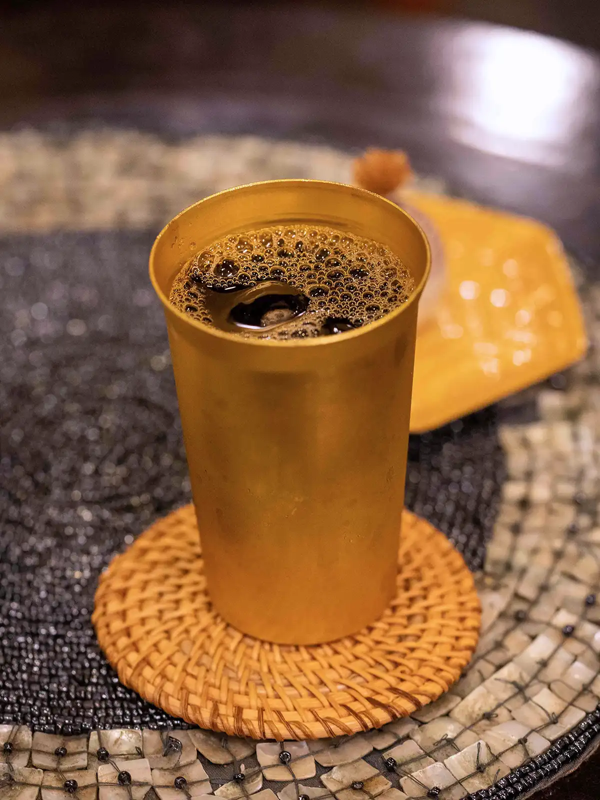 A close-up of a cold coffee in a rustic golden cup on a woven coaster.