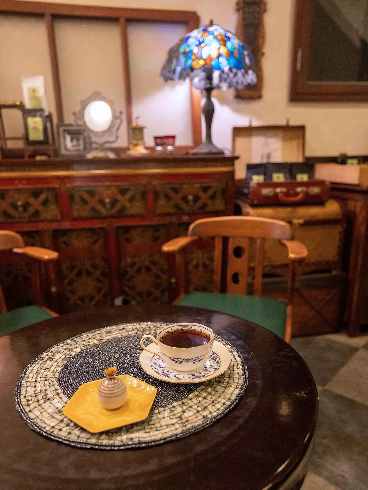 A cup of coffee on a mosaic table with a vintage backdrop and Tiffany lamp.