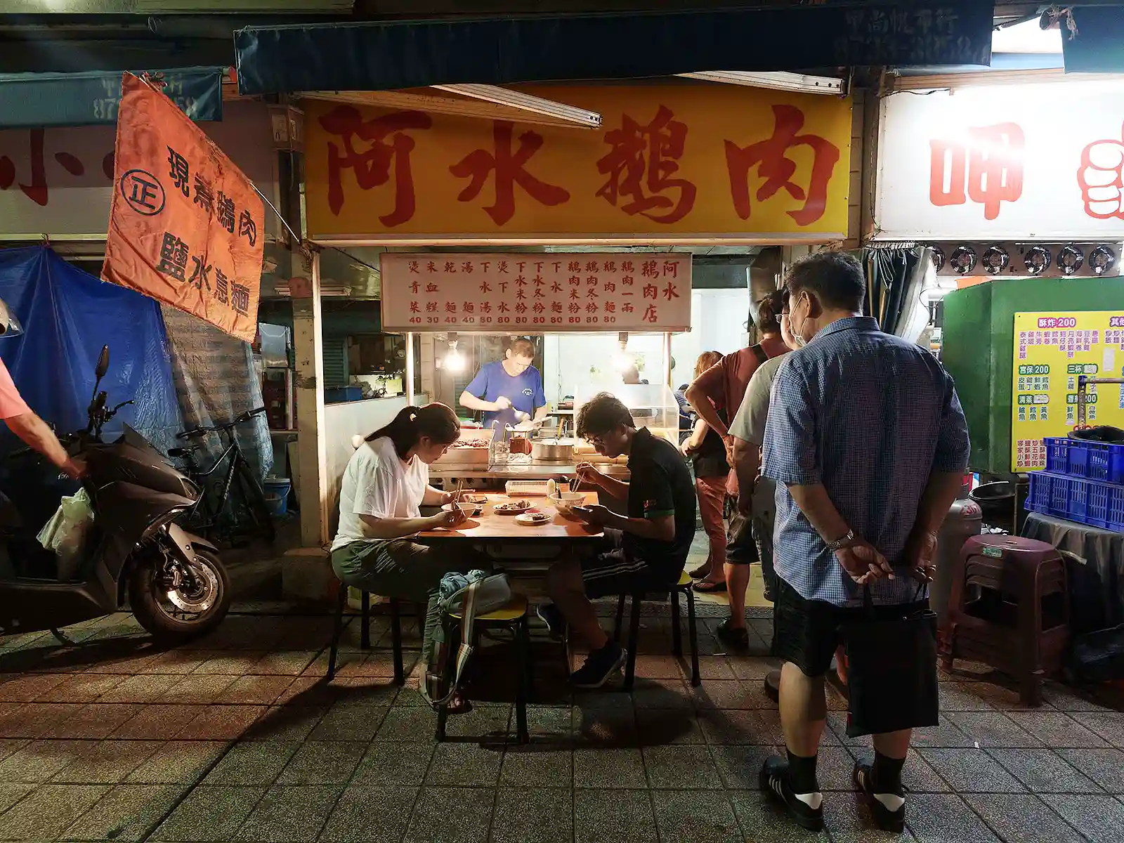 Customers dine at a street-side eatery under glowing signs at a Taipei night market.