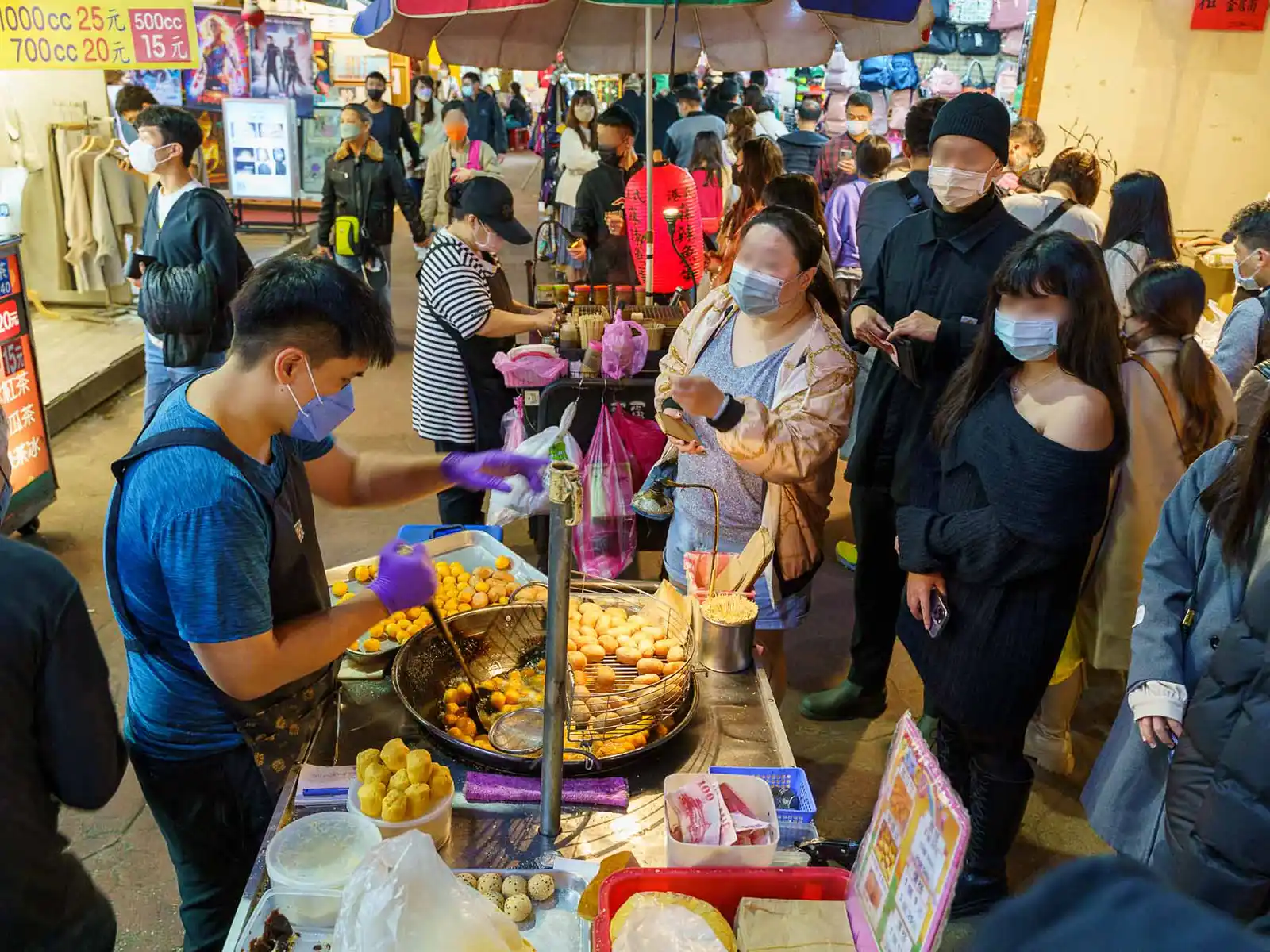 A vendor fries sweet potato balls at his stand while customers queue at a night market.