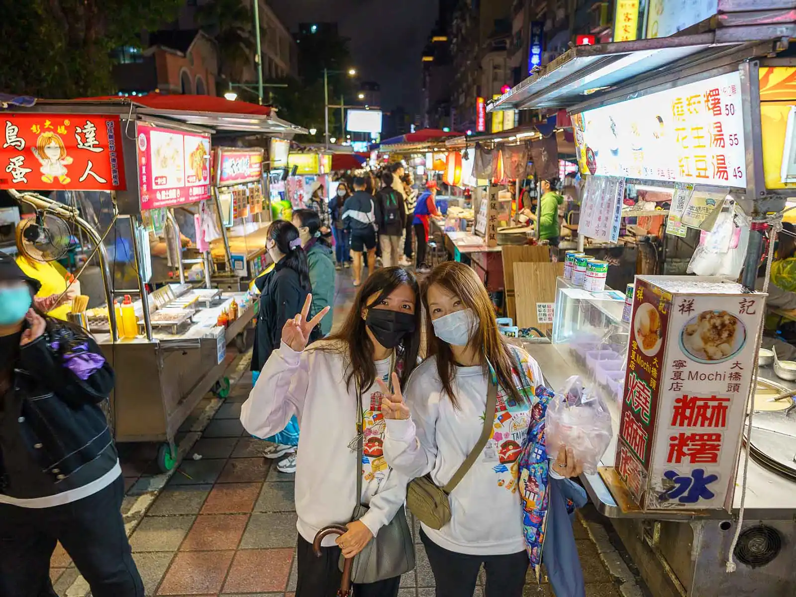 Two friends pose with peace signs at a bustling Taipei night market.