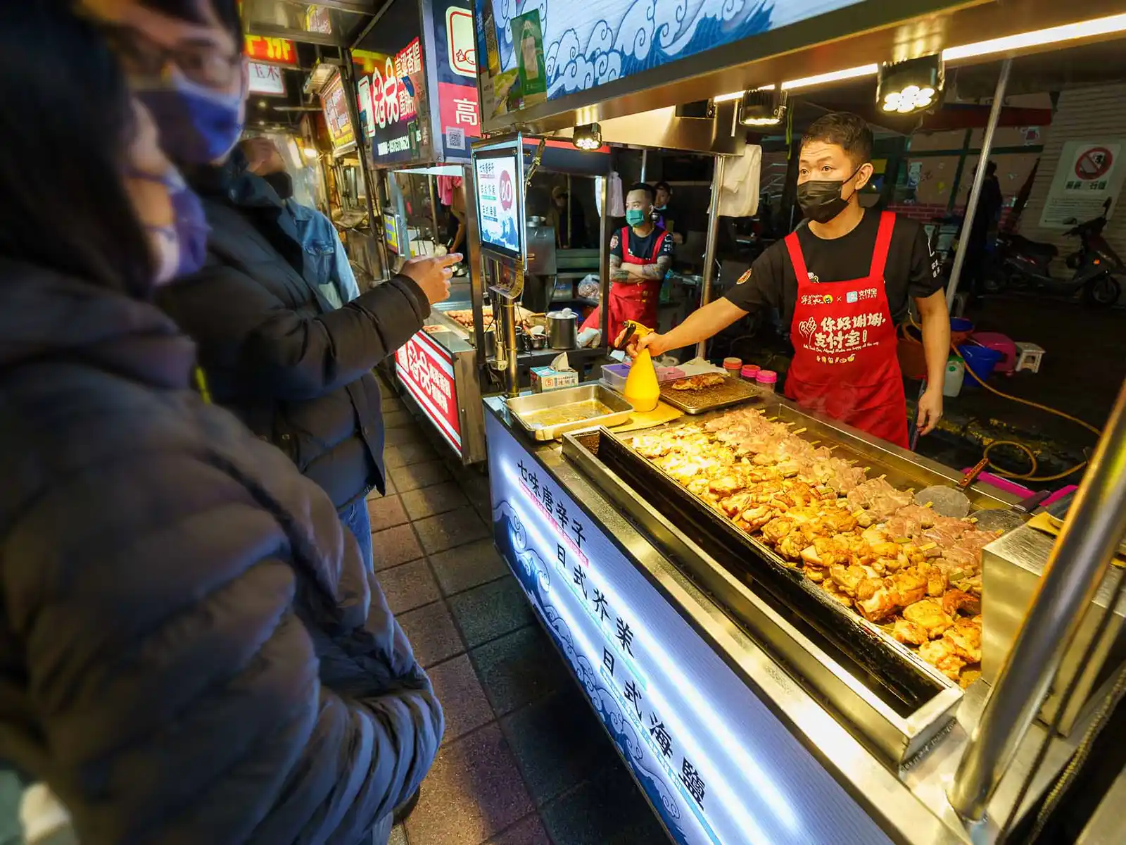 A customer points at a food stall while the vendor sells skewers at a night market.