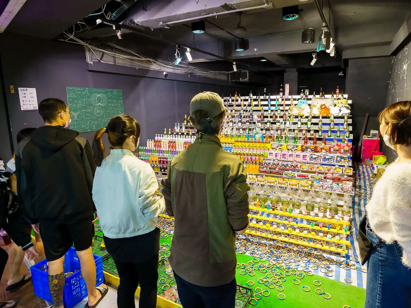 Visitors take turns tossing rings on to rows of bottles at a toss game night market stand.