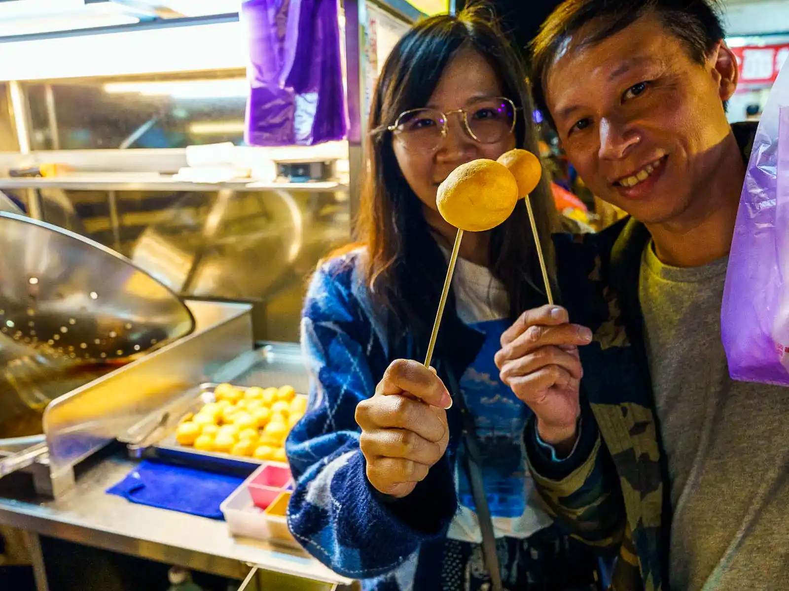 A smiling couple holding up skewered treats at a night market.