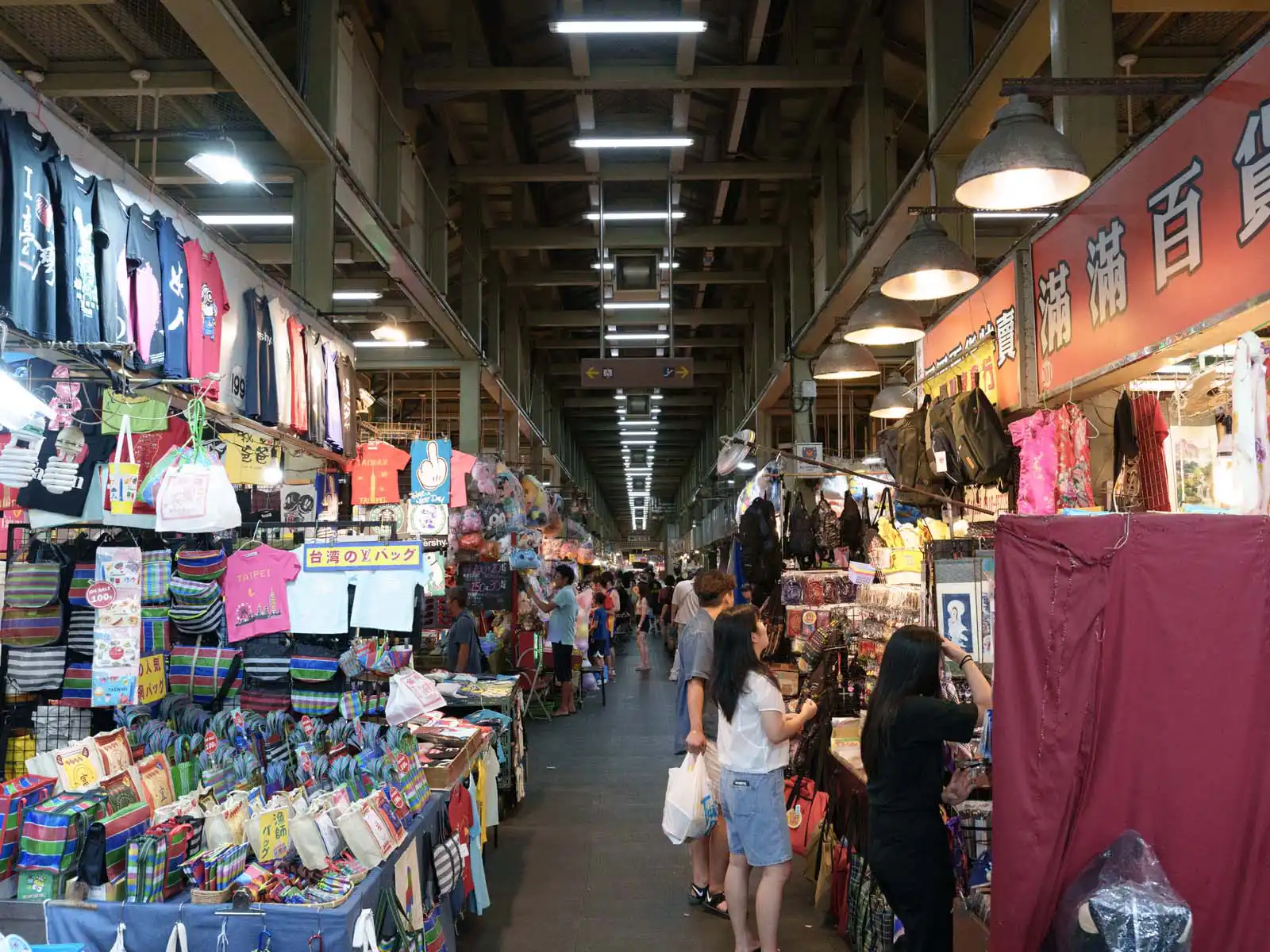 Crowded indoor market scene with clothing stalls lining a long corridor.