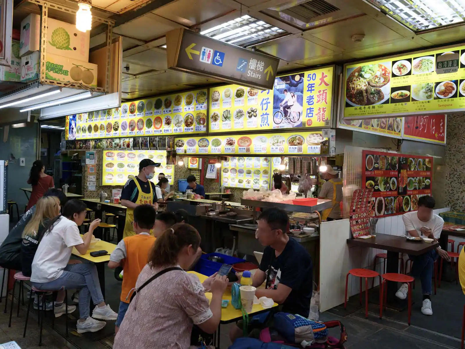 Patrons enjoy meals at a busy food stall with a vibrant menu display in Taipei.